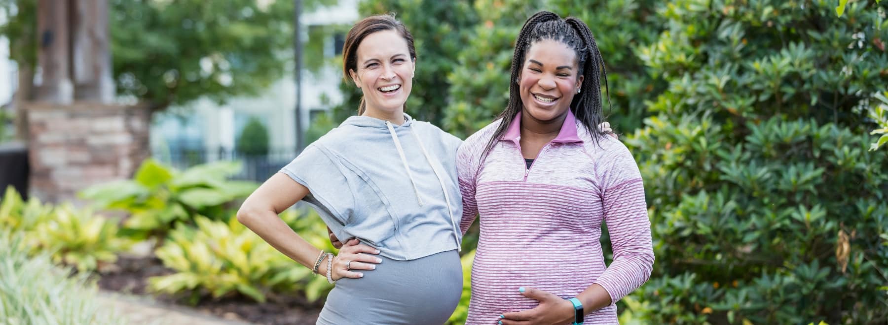 Two pregnant women in exercise clothing smile while posing for a picture in a park